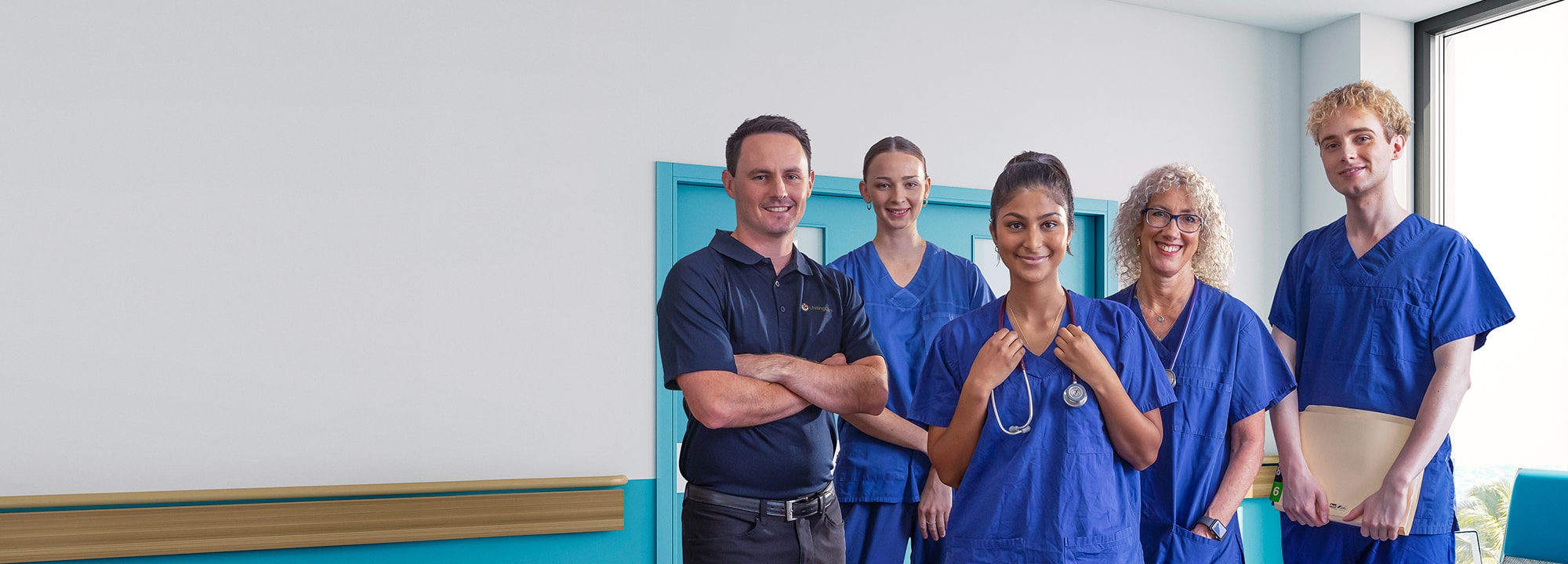 Five hospital workers, some wearing scrubs, stand in a blue hospital corridor, smiling.