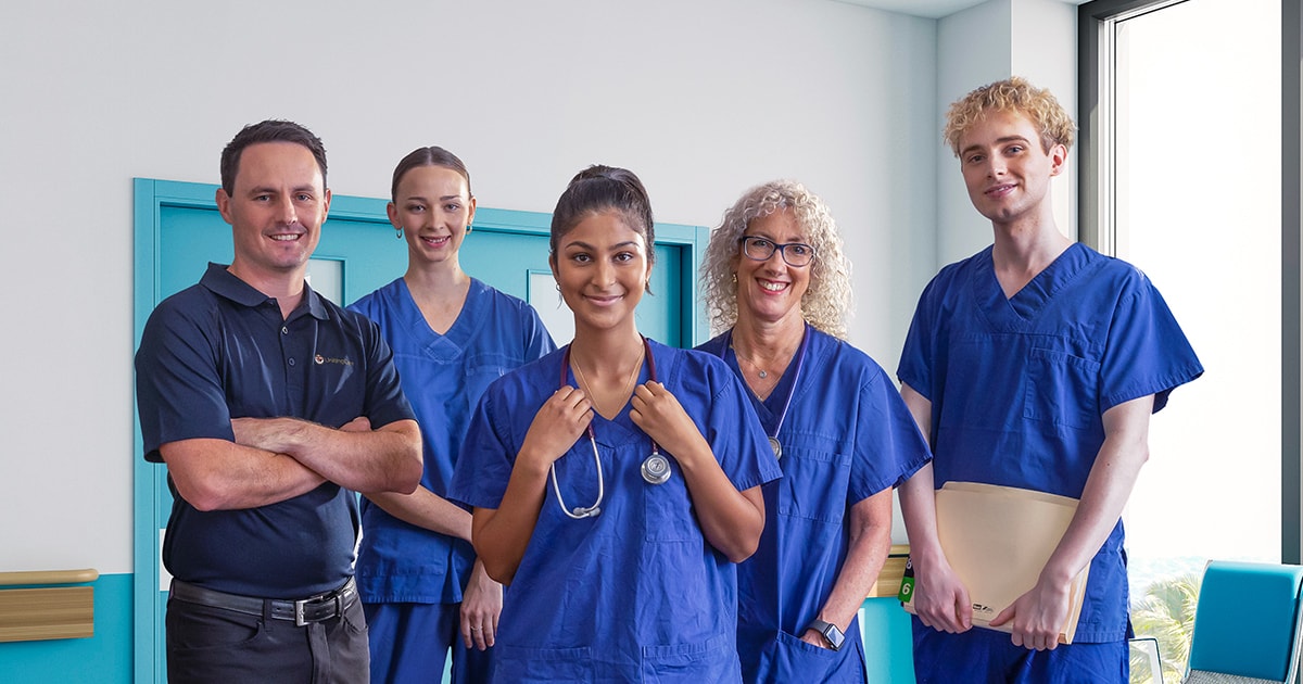 Five hospital workers, some wearing scrubs, stand in a blue hospital corridor, smiling.