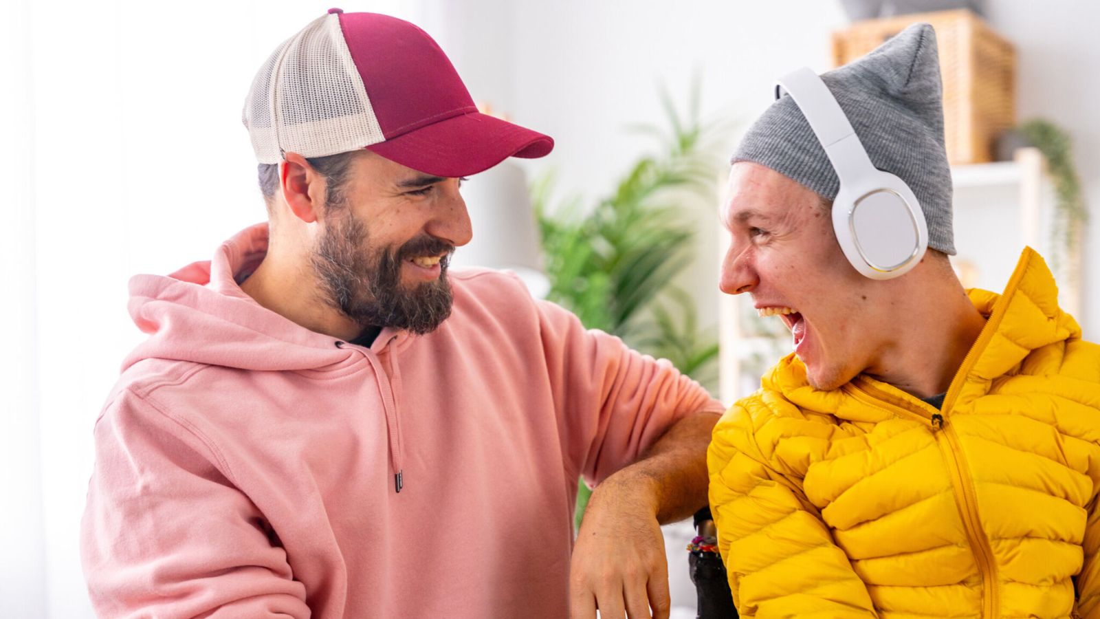 A man in a cap and a pink sweatshirt smiles at a laughing man with disability wearing a gey beanie, white headphones and yellow puffer jacket.