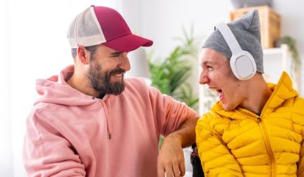 A man in a cap and a pink sweatshirt smiles at a laughing man with disability wearing a gey beanie, white headphones and yellow puffer jacket.