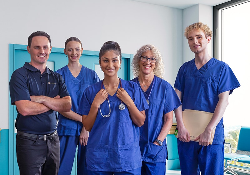 Five hospital workers, some wearing scrubs, stand in a blue hospital corridor, smiling.