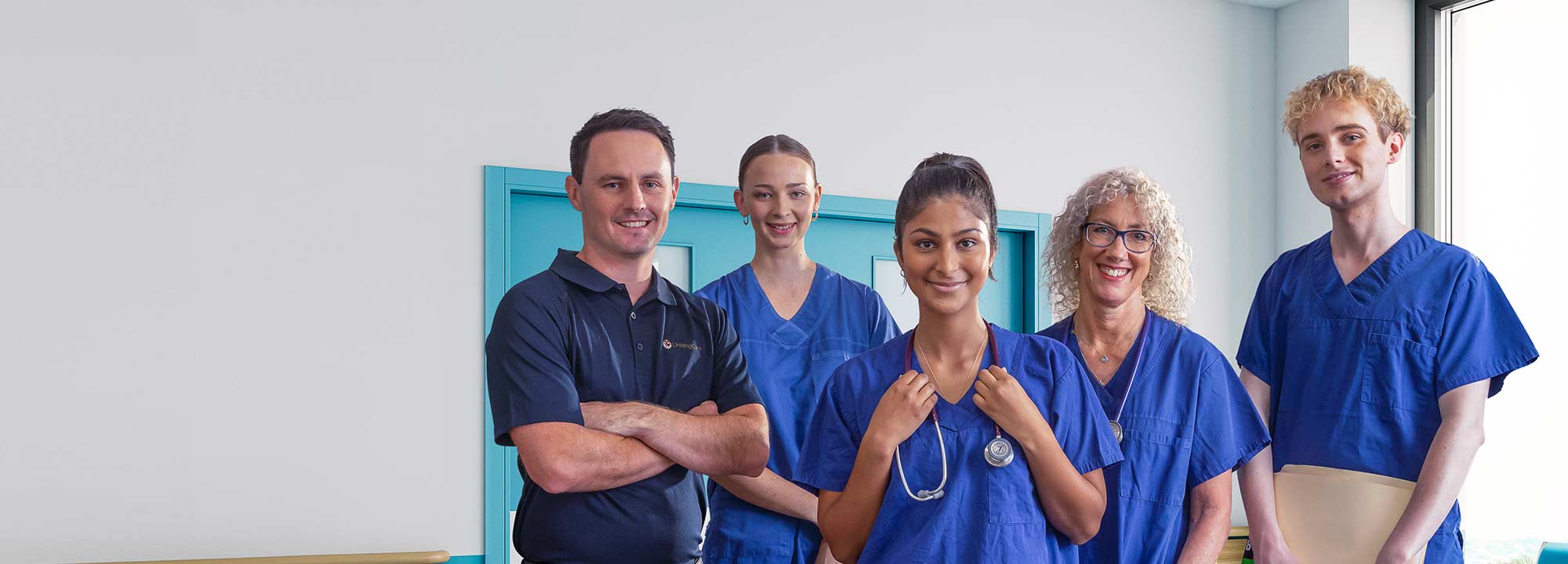 Five hospital workers, some wearing scrubs, stand in a blue hospital corridor, smiling.