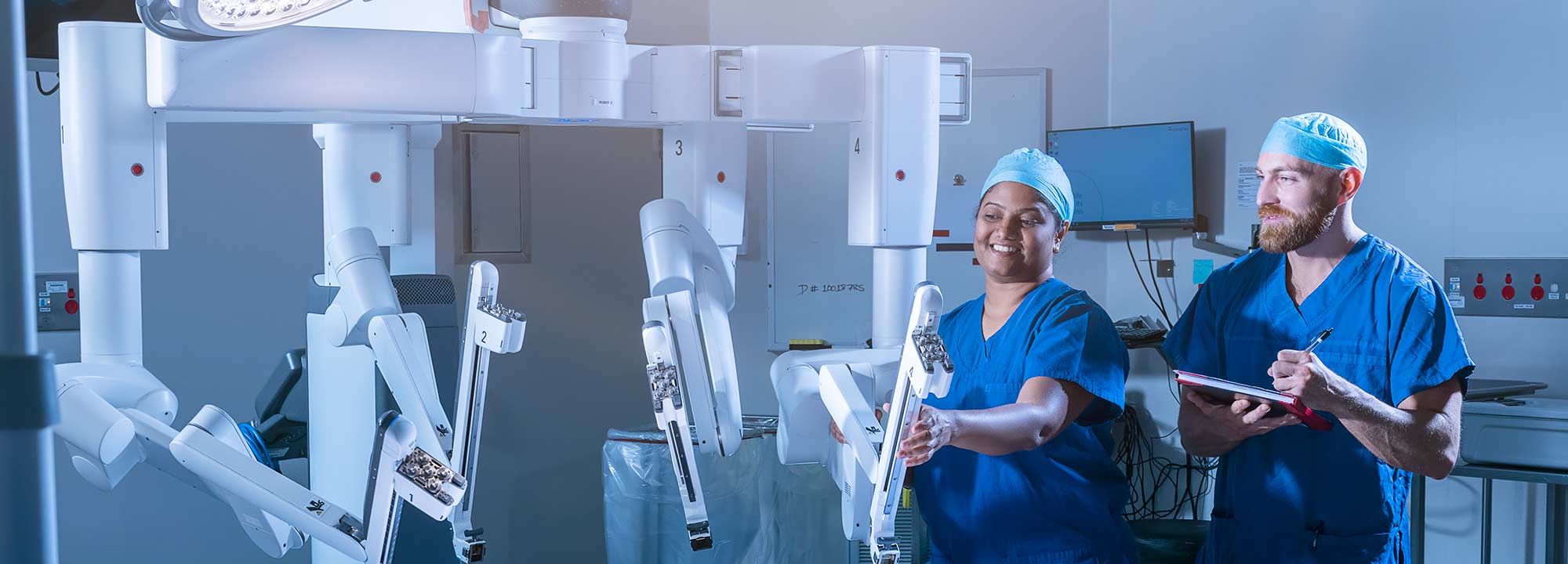 Two hospital workers in blue scrubs stand next to a da Vinci robot in a hospital theatre.