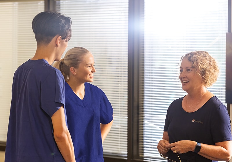 Two hospital workers in blue scrubs talk to a lady near a reception desk.