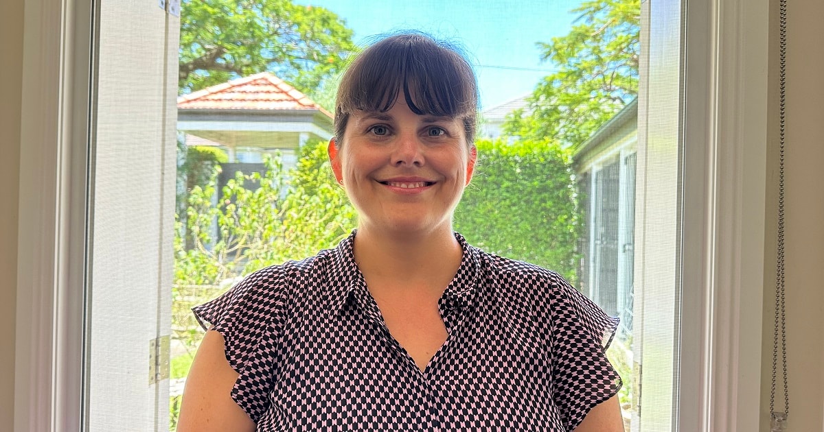 Alicia Jonker Choices Cancer Support A young woman smiling as she stands in front of a screen door, with a sunny backyard behind her