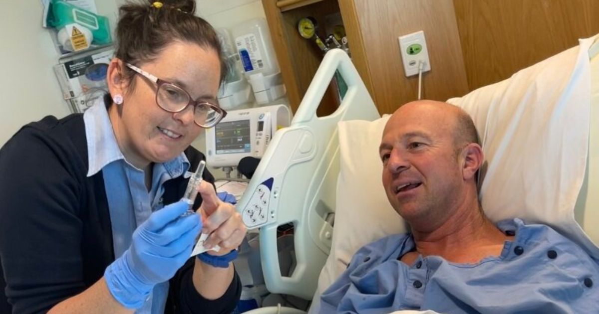 Patient Andrew Parry lies in a hospital bed, talking to a smiling nurse.