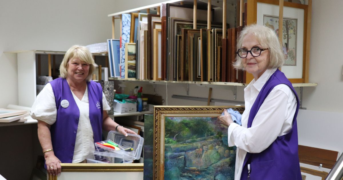 Wesley Hospital volunteers Caroline McCormack and Kathy Praine, wearing purple volunteer vests, stand surrounded by artworks.