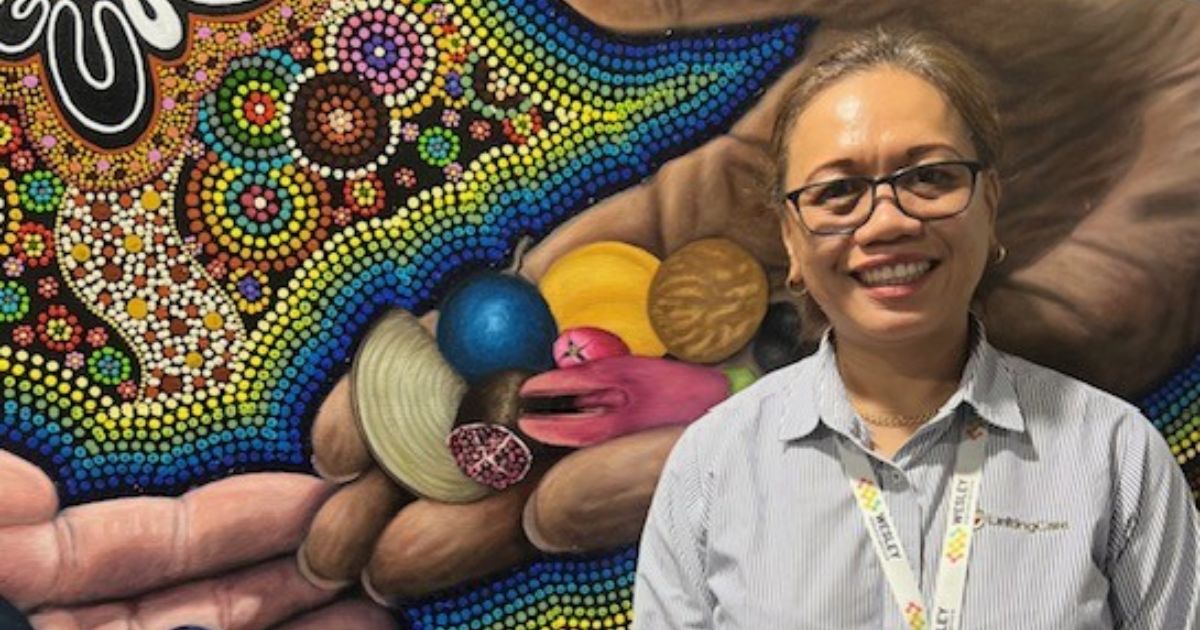 The Wesley Hospital Environmental Auditor and Waste Warrior Mira Fitzgerald stands in front of an Aboriginal artwork which depicts two hands connecting.