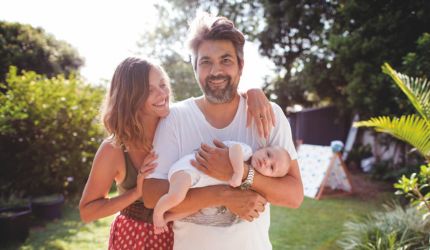 A bearded man holds a young baby in his arms, while his wife hugs him and looks on.