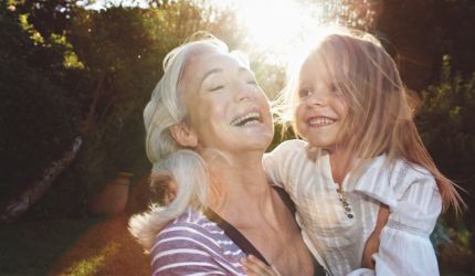 A woman with grey hair holds a young girl in a white shirt as they laugh together in the sunshine.