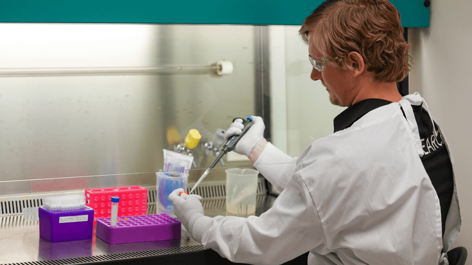 A man in a white coat and safety glasses examines a specimen in a lab.