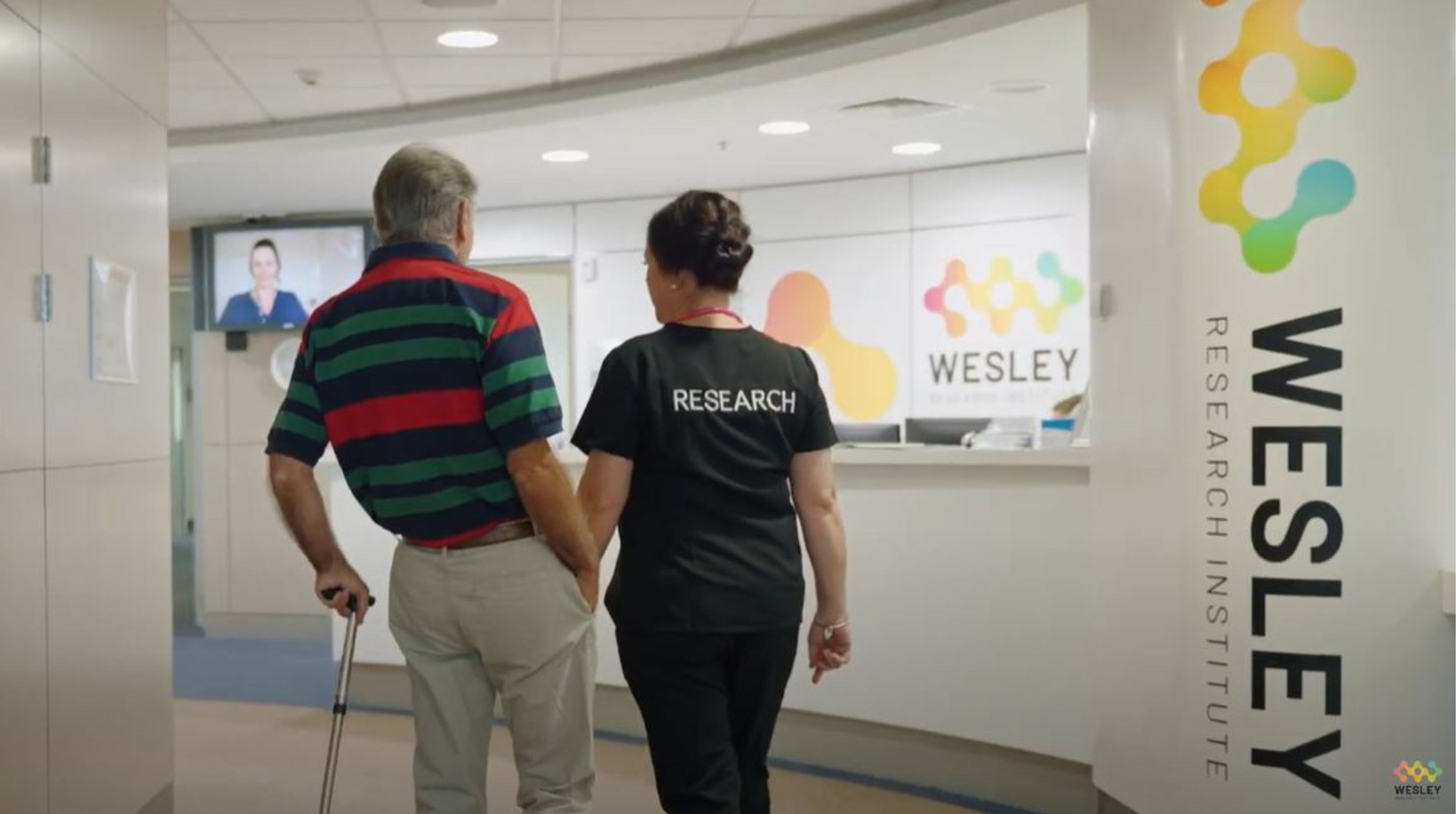 A man with a can and a woman in a Wesley Medical Research T-shirt walk towards the Wesley Research Institute reception desk.