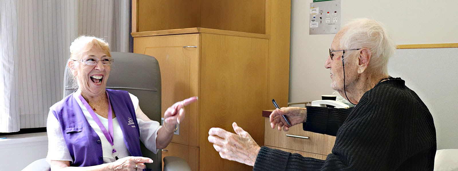 A Wesley Hospital Compassionate Companion shares a laugh with an elderly patient sitting on his hospital bed.