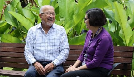 A man in a white checked shirt and a woman in a purple jumper sit on a park bench backed by greenery.