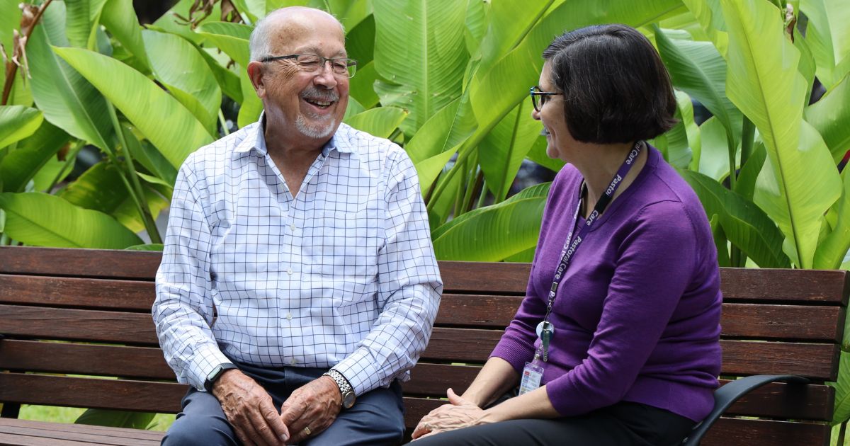 A man in a white checked shirt and a woman in a purple jumper sit on a park bench backed by greenery.