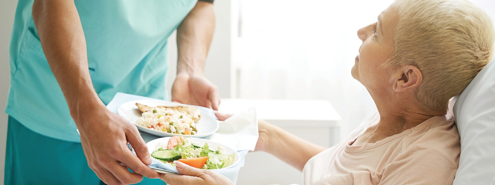 A hospital worker hands a tray of food to a patient sitting in bed.